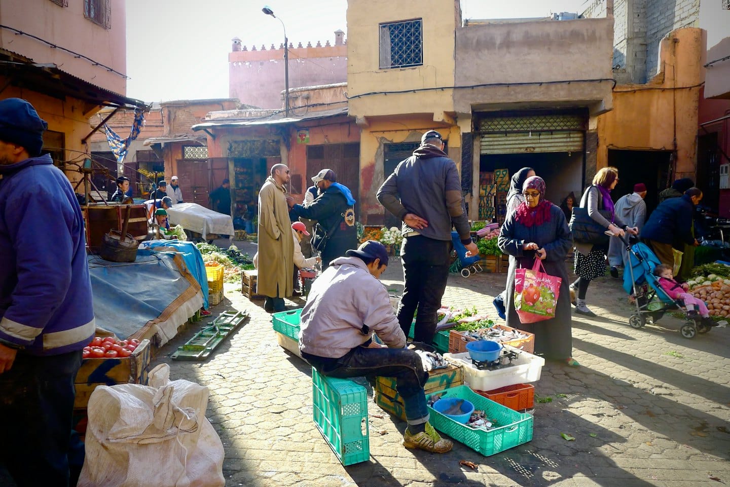 Shoppers browse fresh produce at a busy cobblestone street market.