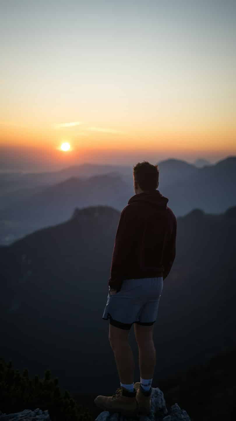 Hiker stands on a rocky mountain peak overlooking layered ranges.
